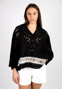 Black poncho with intricate cut-out floral patterns, featuring a white lace trim at the hem. V-neck design and loose sleeves.