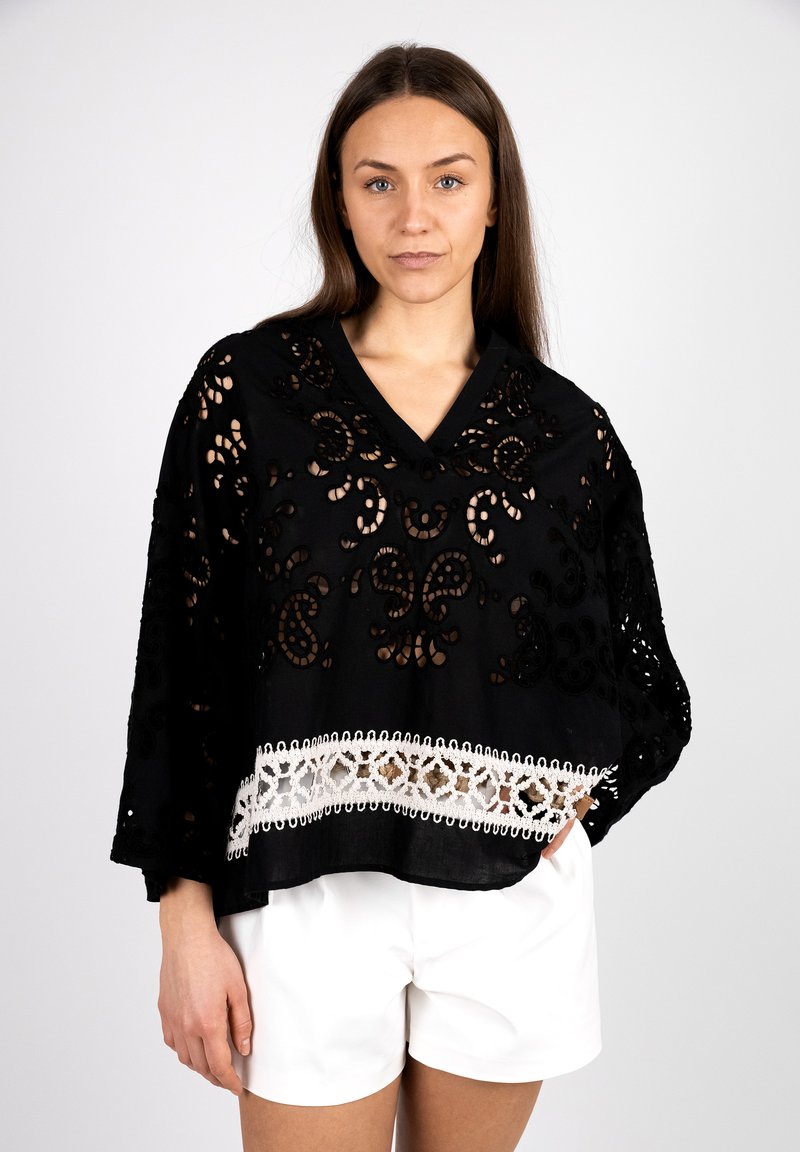 Black poncho with intricate cut-out floral patterns, featuring a white lace trim at the hem. V-neck design and loose sleeves.