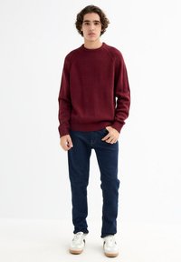 Suéter burdeos con dobladillo acanalado y mangas raglán, combinado con jeans de un azul oscuro y zapatillas blancas con detalles en negro.