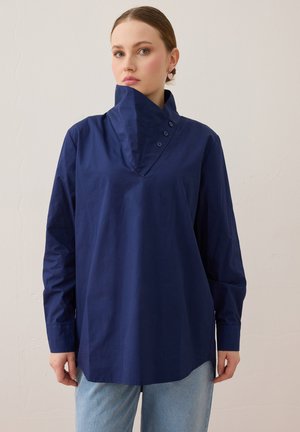 Kvinde iført en løs mørkeblå skjorte med knapper og lange ærmer samt lyseblå jeans mod en ensfarvet baggrund.