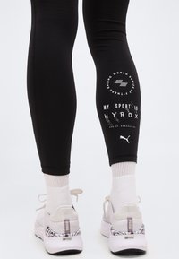 Πόδια που φοράνε μαύρο κολάν με το κείμενο «MY SPORT IS HYROX» και λογότυπα, σε συνδυασμό με λευκά αθλητικά παπούτσια Puma και λευκές κάλτσες σε απλό φόντο.