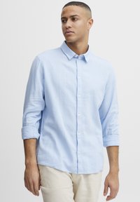 Lys blå, langermet skjorte med knapper laget av lin, med en klassisk krage og oppbrettede ermer. Hvite knapper foran.