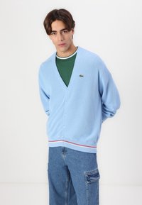 Cardigan azzurro chiaro con scollo a V, dotato di sei bottoni e orlo a righe rosse e bianche. Include un piccolo logo Lacoste verde.