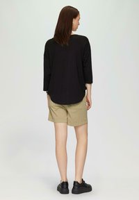 Czarny top z trzema kwartami rękawów w połączeniu z beżowymi szortami. Czarne sneakersy. Gładki materiał, luźny krój, zaokrąglony dół i minimalistyczny design.