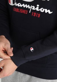 Ręce regulujące mankiet czarnej bluzy z napisem "Champion Established 1919" oraz małym logiem Champion na rękawie.