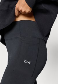 Μαύρο αθλητικό κολάν με λεία υφή, διαθέτει ψηλή μέση και διακριτικό λευκό λογότυπο στο πλάι. Είναι ορατά ραφές στο σχεδια�σμό.