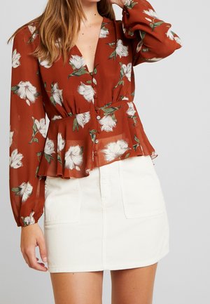 Femme portant un chemisier à manches longues couleur rouille imprimé de fleurs blanches avec des boutons, associé à une mini-jupe blanche avec poches devant.