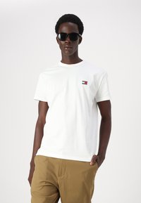 Tricou alb din bumbac cu mâneci scurte, având un mic logo roșu, alb și albastru pe partea stângă a pieptului. Purtați cu pantaloni bej deschis.