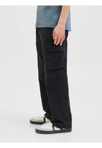 Pantalons cargo noirs à coupe ample, dotés de poches latérales et d'une fermeture à rabat. Portés avec des baskets blanches et grises.