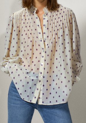 Woman wearing loose cream blouse with purple polka dots, buttoned front, and blue jeans, hands resting in pockets.