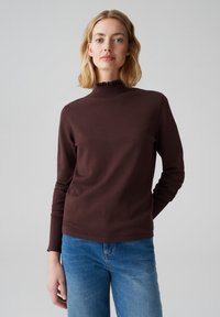 Braunes Langarmoberteil mit hohem Rüschensaumkragen, begleitet von blauen Jeans. Der Stoff wirkt glatt und enganliegend.