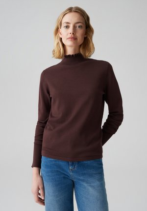 Top lungo a maniche con colletto alto arricciato di colore marrone, accompagnato da jeans blu. Il tessuto sembra liscio e aderente.