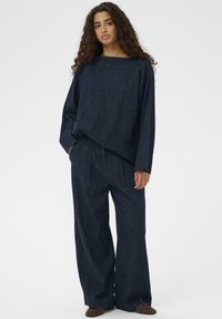 Žena s dlhými kučeravými vlasmi, oblečená v voľnej tmavomodrej rifľovej blúzke a širokých nohaviciach, stojaca s jednou rukou v vrecku na bielom pozadí.