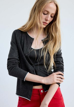 Jeune femme aux longs cheveux blonds portant un cardigan gris foncé et un haut noir, ajustant sa manche, et un pantalon rouge sur un fond uni.