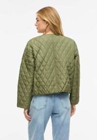 Moteris su ilgais blondinais plaukais, dėvinti alyvuogių žalios spalvos siuvinėtą striukę ir šviesiai mėlynas džinsines kelnes, atsukta nugara priešais paprastą baltą foną.