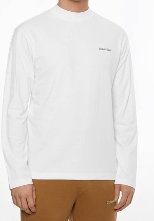 Mężczyzna noszący prostą białą koszulę z długim rękawem Calvin Klein oraz brązowe spodnie z logo Calvin Klein na kieszeni.