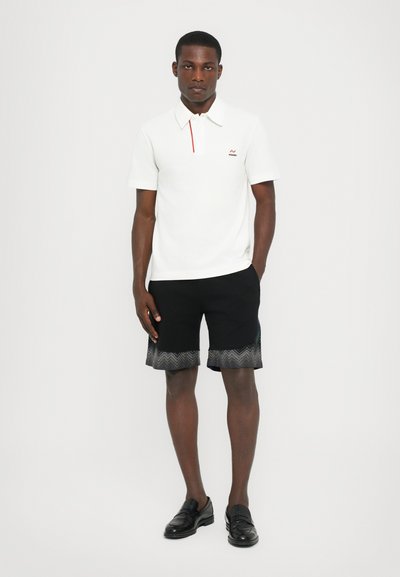 Camisa polo branca com um detalhe vermelho, combinada com calções pretos com um padrão zig-zag na bainha. O modelo usa sapatos mocassins pretos.