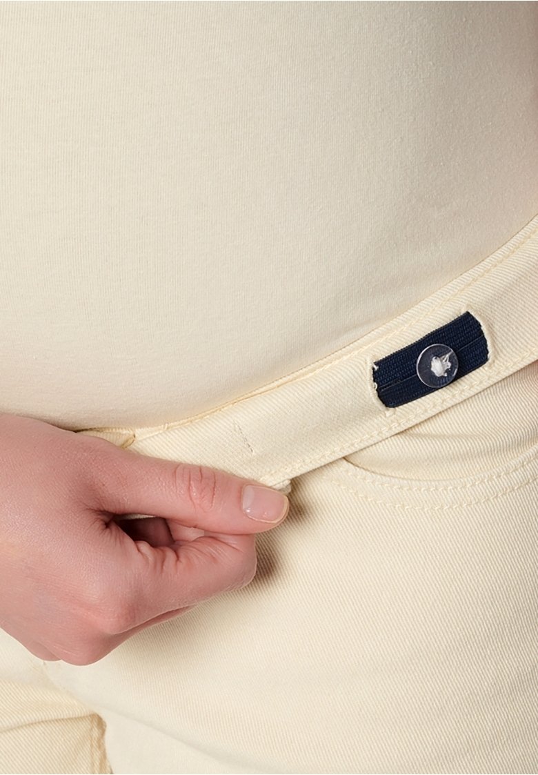 Pantalón de tela beige texturizada con cierre de botón y etiqueta de acento azul marino. La mano se muestra sujetando la cintura para ajustar.