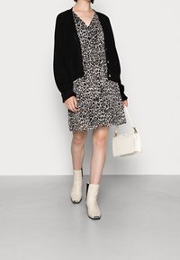 Schwarze Strickjacke über einem beigen Kleid mit Leopardenmuster und Bindegürtel, kombiniert mit cremefarbenen Stiefeletten und einer kleinen weißen Handtasche.