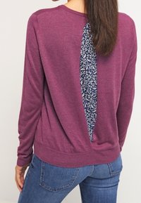 Fioletowy sweter z długim rękawem z wstawką z wzorzystego niebieskiego materiału na plecach, w parze z niebieskimi dżinsami o luźnym kroju.