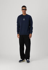 Sudadera navy con un logo triangular, combinada con pantalones negros holgados y zapatillas azules. El modelo lleva gafas de sol oscuras.