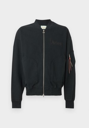 Homme aux cheveux bouclés portant des lunettes de soleil noires, une veste bomber noire sur un t-shirt blanc avec un motif graphique, un pantalon noir à jambes larges et des bottes couleur beige, debout.