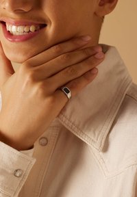 Zilveren ring met een rechthoekige zwarte steen, gedragen aan een vinger, tegen een achtergrond van een lichtgekleurde stoffen kraag en de huidtextuur van de hand.