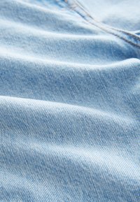 Lyseblåt denimmateriale med en blød tekstur, der har et diskret fiskebenmønster og kontrasterende orange syninger langs sømmene.