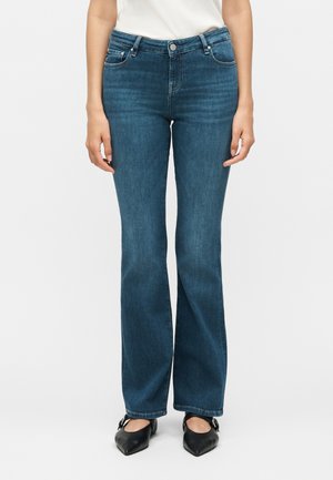 Nő magasderekú kék bővülő szárú farmert és fekete hegyes orrú balerinacipőt visel, lazán lógó kezekkel áll.