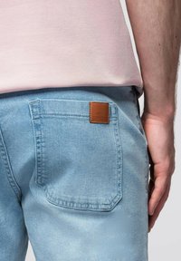 Světle modré džíny z denimu s zadní kapsou a hnědou koženou nášivkou. Tkanina se zdá být měkká a s jemnou texturou. Částečně je vidět růžová košile.