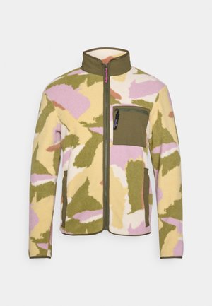 Flis jakna u žutoj, zelenoj i ružičastoj maskirnoj uzorci. Ima visok ovratnik, patentni zatvarač pune duljine i prsni džep s patentnim zatvaračem.