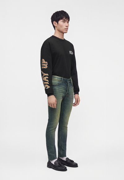 Jeune homme debout portant une chemise noire à manches longues avec un texte sur la manche, un jean vert délavé, des chaussettes blanches et des mocassins noirs à pampilles.