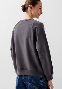 Felpa grigia con colletto tondo e polsini a costine. Il tessuto sembra testurizzato, con una vestibilità comoda, abbinata a pantaloni di jeans blu.