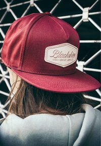 Cappellino da baseball bordeaux in tessuto con visiera piatta, caratterizzato da un patch logo ricamato color crema e dettagli di cucitura visibili.