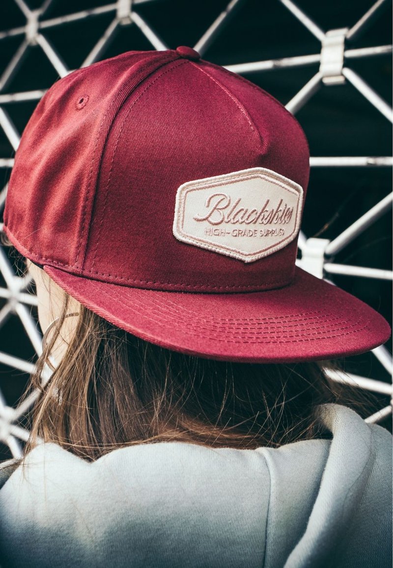 Cappellino da baseball bordeaux in tessuto con visiera piatta, caratterizzato da un patch logo ricamato color crema e dettagli di cucitura visibili.