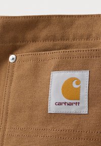 Primo piano di un tessuto marrone con una texture, con una toppa quadrata cucita di colore bianco che presenta il logo giallo di Carhartt e il nome del marchio in rosso.