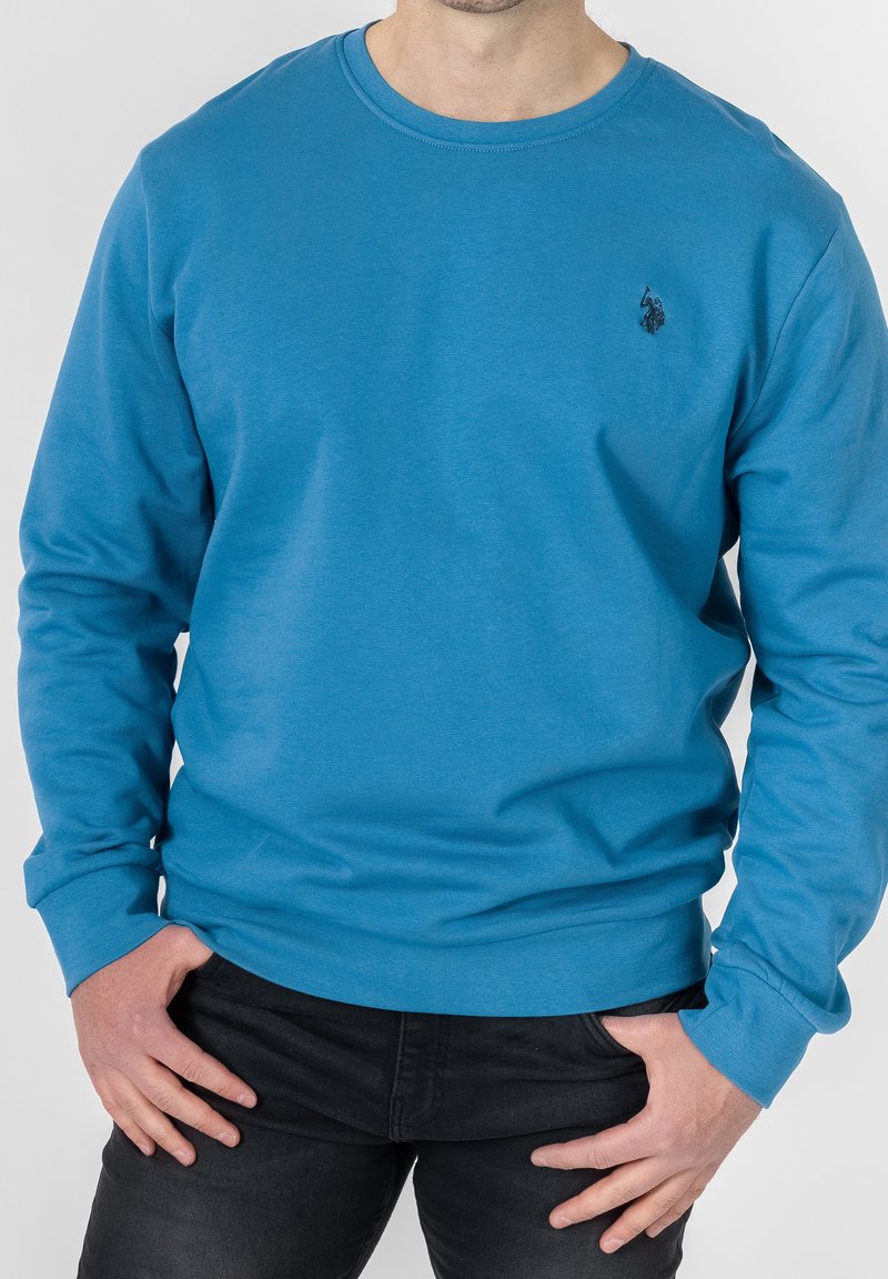 Blå långärmad sweatshirt med rund halsringning, tillverkad av mjukt tyg, med en liten svart emblem på vänster bröst och ribbade ärmslut.