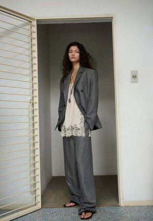 Vrouw met krullend haar staat in het deurkozijn, gekleed in een oversized grijs pak, crème kanten top, zwarte slippers en een lange ketting met hanger.