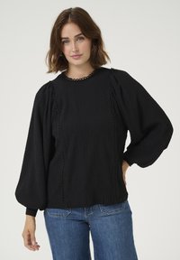 Black blouse with textured fabric, round neckline, and puffed sleeves. Features contrasting stitched detailing along edges and is paired with denim jeans.