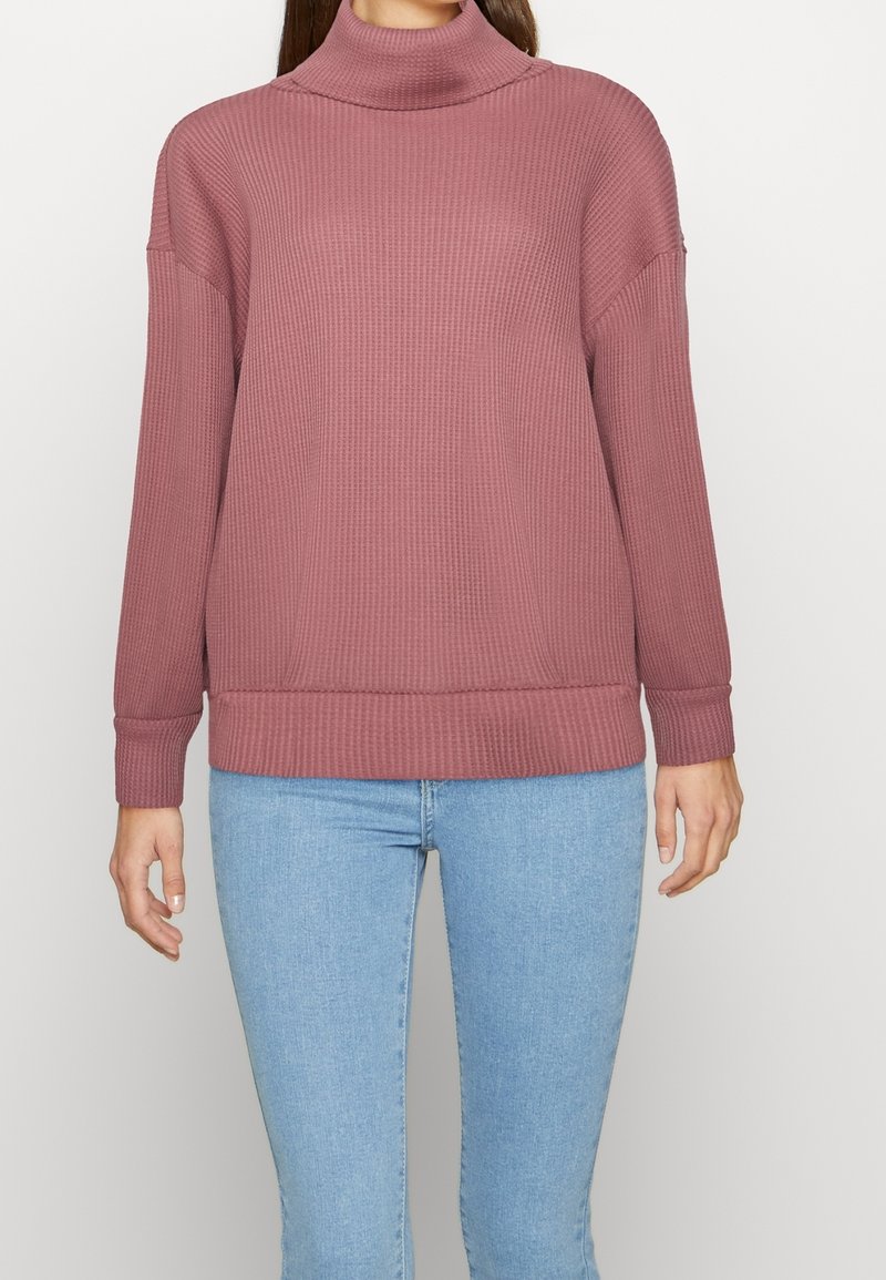 Femme portant un pull à col roulé côtelé rose poussière et un jean bleu clair sur un fond uni.
