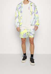 Kolorowa kurtka i szorty z graficznym wzorem zielonych i fioletowych kształtów, połączone z beżową bluzą i czarnymi tenisówkami.