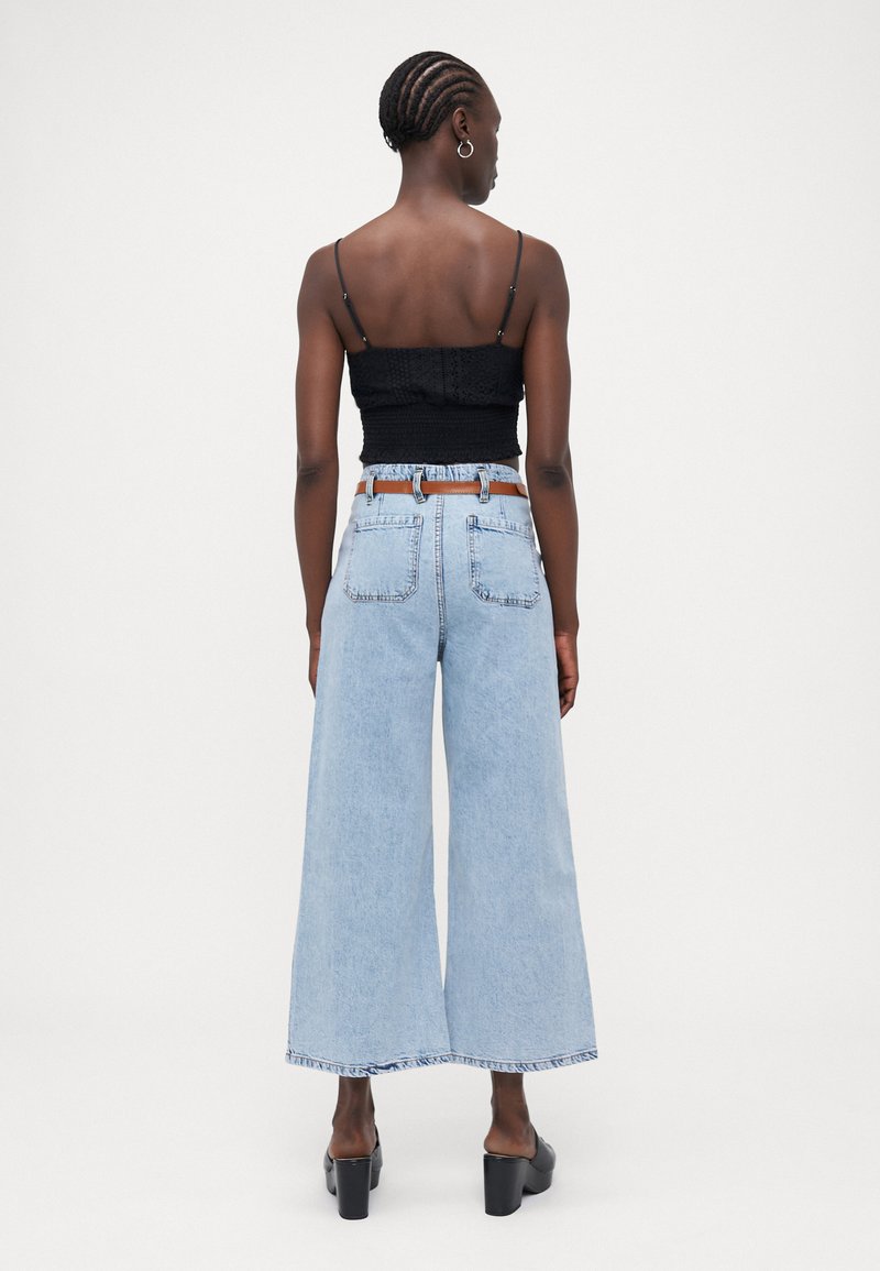 Jeans larghi blu chiaro con orlo grezzo, abbinati a un top corto nero con arricciature. L'outfit è completato da una cintura beige e tacchi neri chunky.