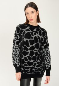 Giovane donna che indossa un pullover morbido a motivi neri e grigi e pantaloni di pelle neri, che si trova in piedi su uno sfondo chiaro e uniforme.