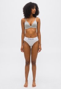 Femme debout pieds nus devant un fond blanc, portant un soutien-gorge triangle gris et une culotte blanche avec le logo Polo.