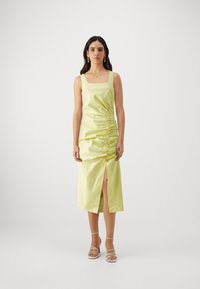 Robe midi jaune à encolure carrée, plissée sur les côtés et dotée d'une fente avant, fabriquée en tissu lisse. Modèle portant des talons à lanières.