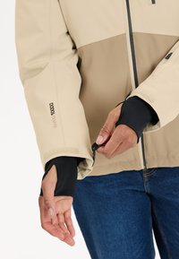 Beige en bruine jack met een rits aan de voorkant, elastische zwarte manchetten en verstelbare klittenband tabs. Het materiaal lijkt glad met minimale textuur.