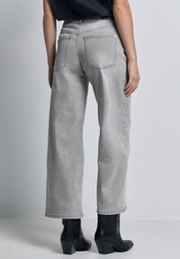 Persona che indossa jeans grigi chiari a gamba larga e stivaletti neri con tacco, mostrata di spalle su uno sfondo semplice.
