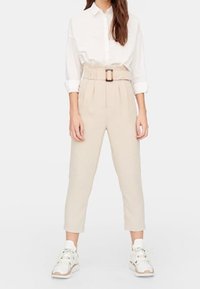 Béžové vysoké kalhoty s páskem, sklady v přední části, zúžené nohavice, spárované s bílou košilí na knoflíky a bílými teniskami.