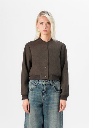 Jeune femme aux cheveux blonds et raides porte une veste boutonnée marron foncé et un jean bleu ample, debout devant un fond blanc uni.