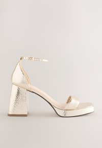 Arany platformok texturált felülettel, bokapánttal és széles, blokk sarkú sarokkal. A dizájn egyenes elejű és lekerekített orrú.